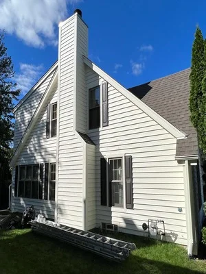 White painted house exterior with vinyl siding, brick chimney, and gable roof against blue sky