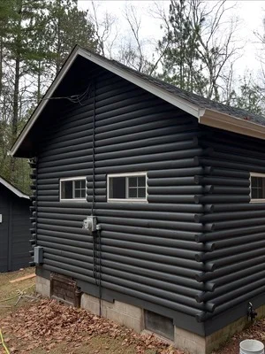 Black painted log cabin exterior with windows and doors, surrounded by trees and mulch landscaping